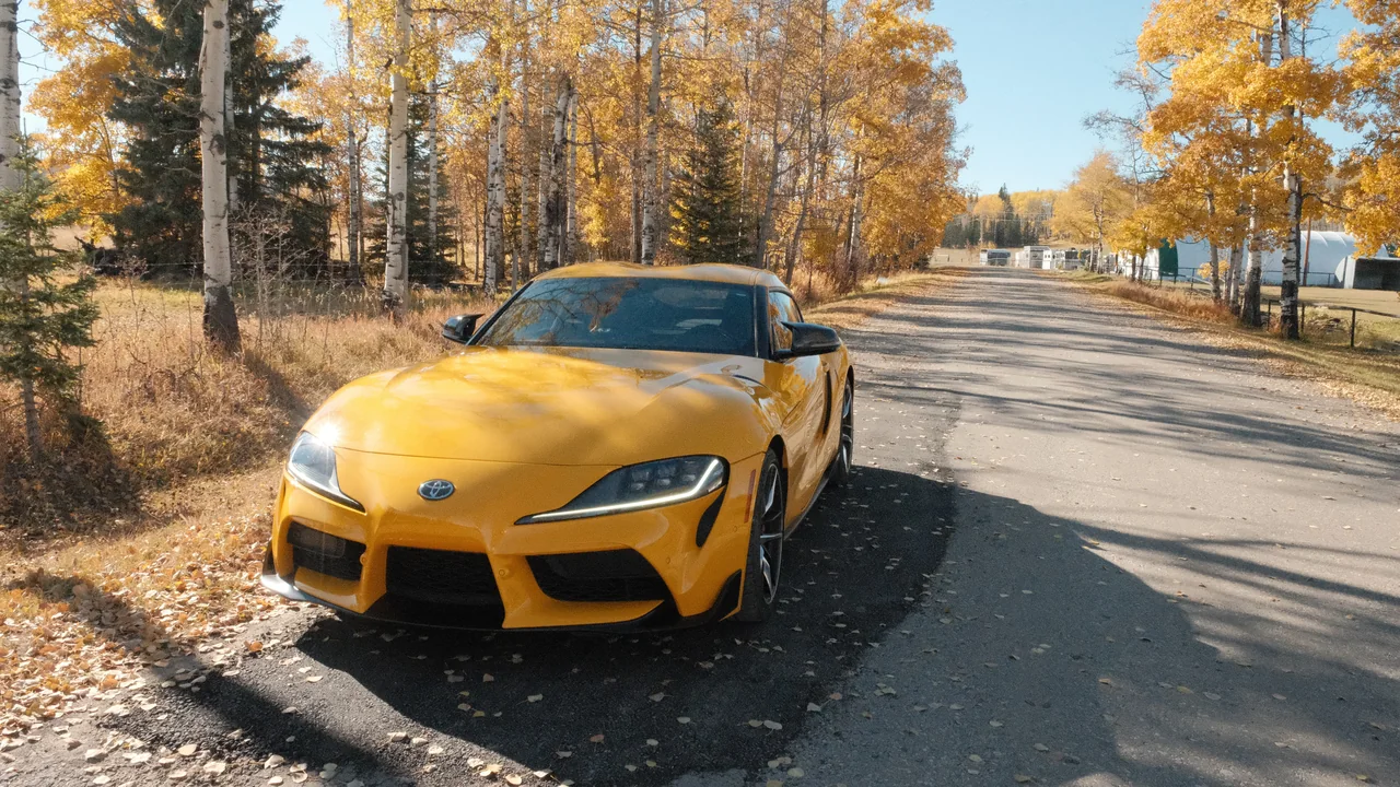 A yellow Toyota Supra parked on a quiet road among golden autumn trees